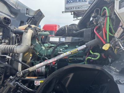 Engine bay of a 2023 Volvo VNL64T760 truck showing engine components, hoses, wiring, and labels visible under the hood.