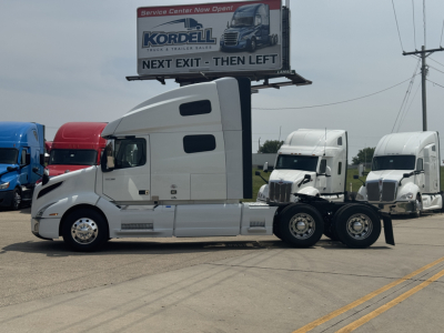 Truck tractor side view showing 2023 Volvo VNL64T760 sleeper cab with quad rear axles, white paint, chrome fuel tanks, and dealership billboard in background.