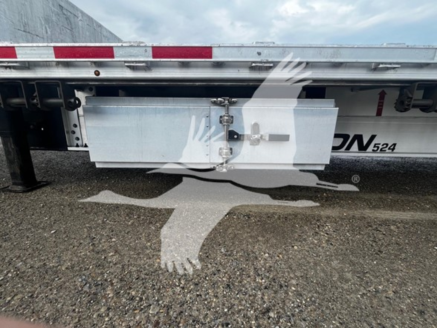 Trailer side view showing aluminum toolbox mounted beneath drop deck with latch and hydraulic lines; partial model label 'ON 524' and reflective tape along top edge.