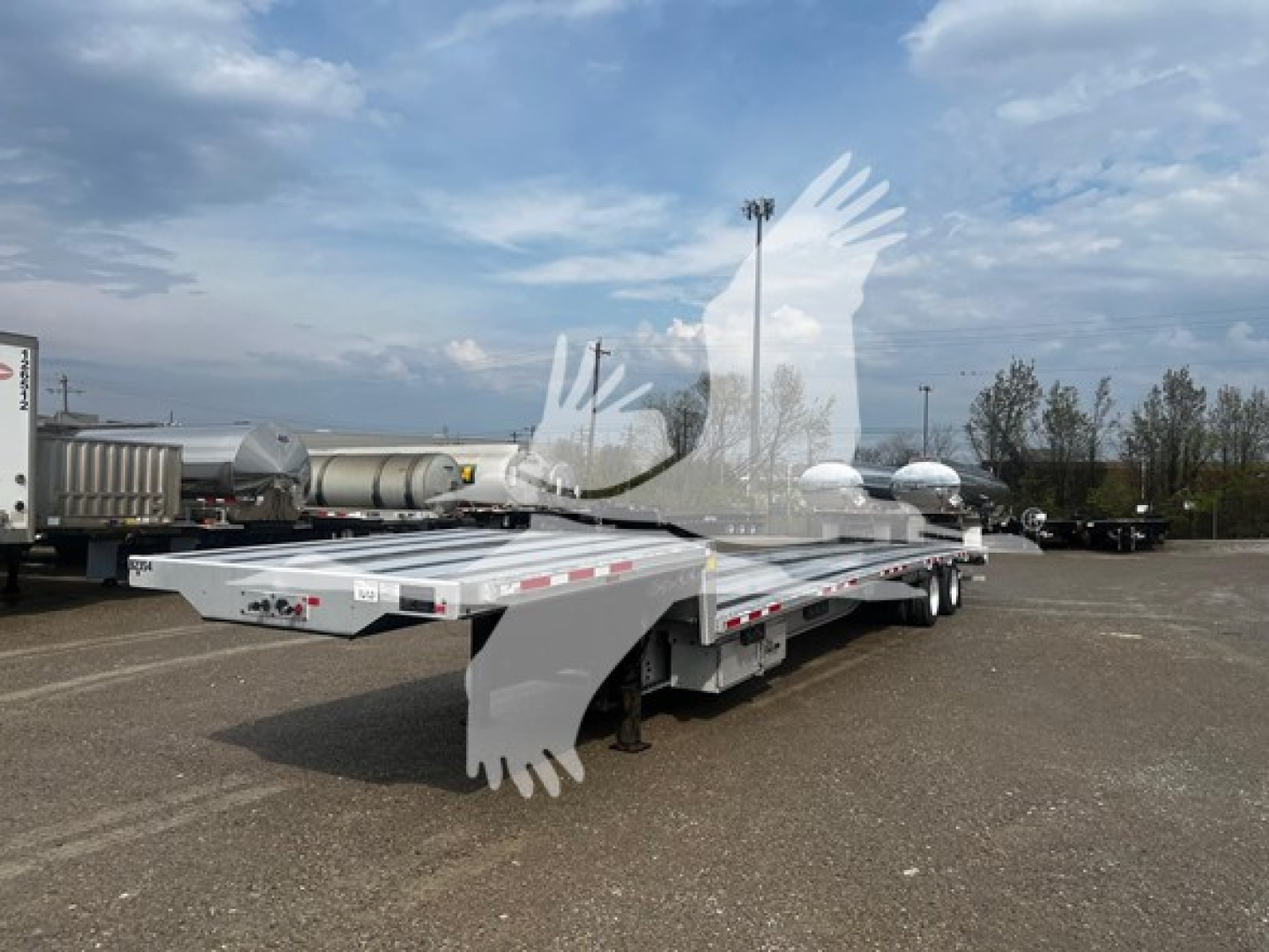 Transcraft aluminum drop deck trailer in a yard showing dual axles and a long flat deck with reflective safety tape.