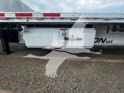 Trailer side view showing aluminum toolbox mounted beneath drop deck with latch and hydraulic lines; partial model label 'ON 524' and reflective tape along top edge.
