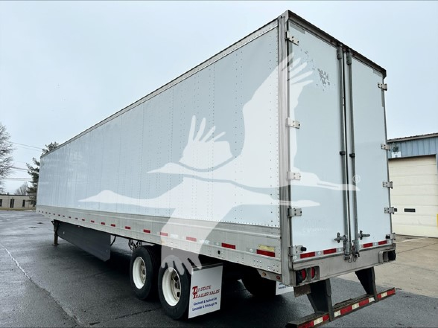 Trailer side view showing a large aluminum box trailer with dual rear doors, reflectors, and a white graphic watermark of geese. Visible text on mud flap and markings include small dealership label.