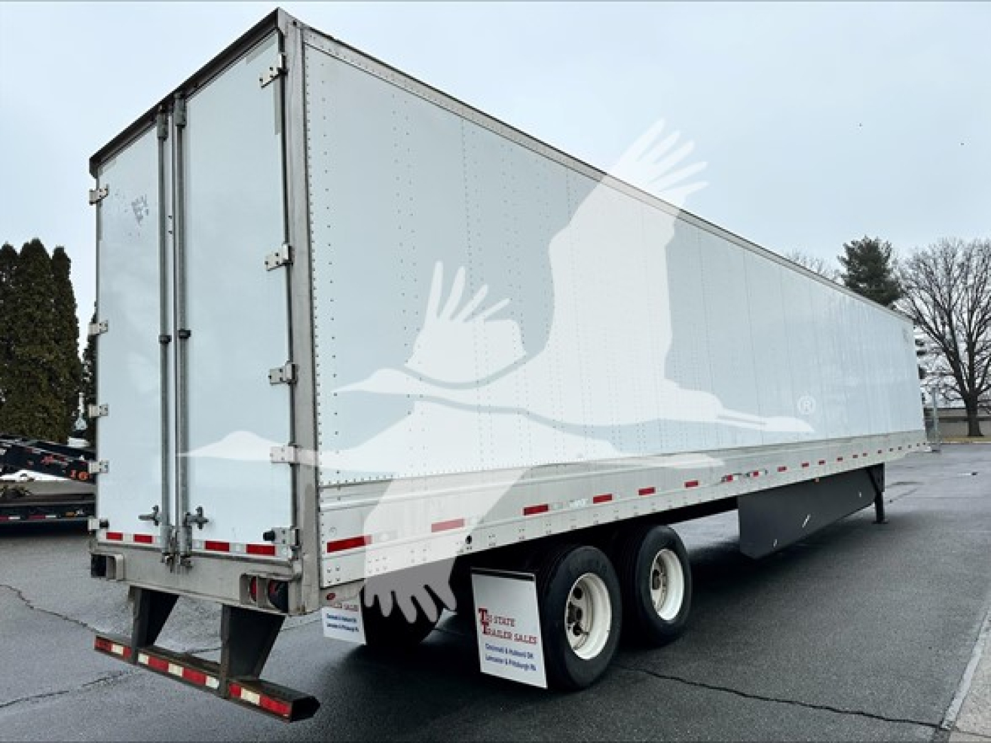 Utility 4000D-X trailer in a lot, side view featuring a white/gray exterior with a faded graphic of geese mid-flight on the side. Dual rear doors with hinges, triple-axle at rear, mudflaps, and reflective safety tape along lower edge.