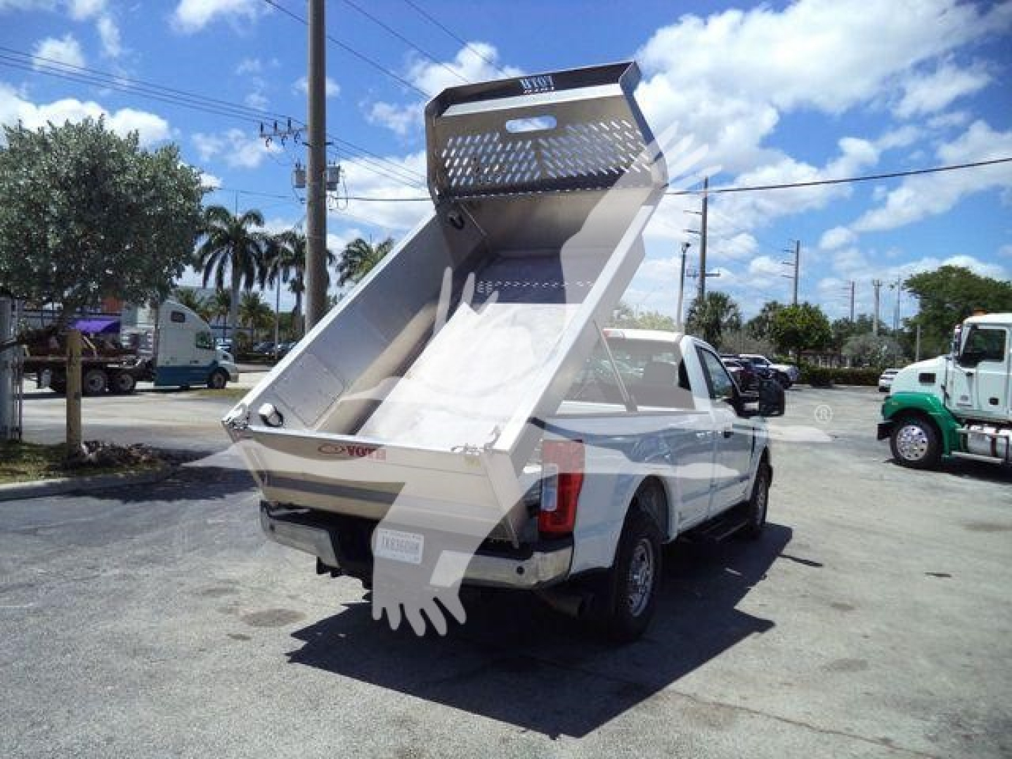 View of a white Volvo 8 ft dump bed truck with a raised aluminum dump bed and rear tailgate. Vehicles and palm trees in the background.