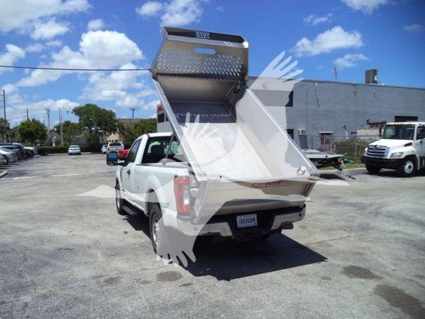 Truck bed with aluminum dump body raised, rear view showing tailgate and hydraulic mechanism. Board reads Volvo? top edge appears with diamond plate panel. Plate on back shows DEF? No legible text. The bed is 8 ft wide.