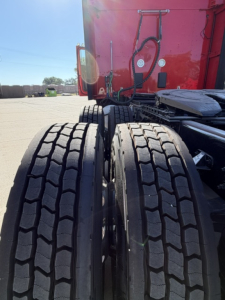 Kenworth truck rear view showing dual rear tires with tread pattern and red cab/tractor in background.