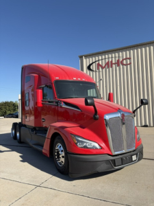 Kenworth T680 semi-truck in red with large chrome grille, front view, in an outdoor lot beside a building with MHC logo.