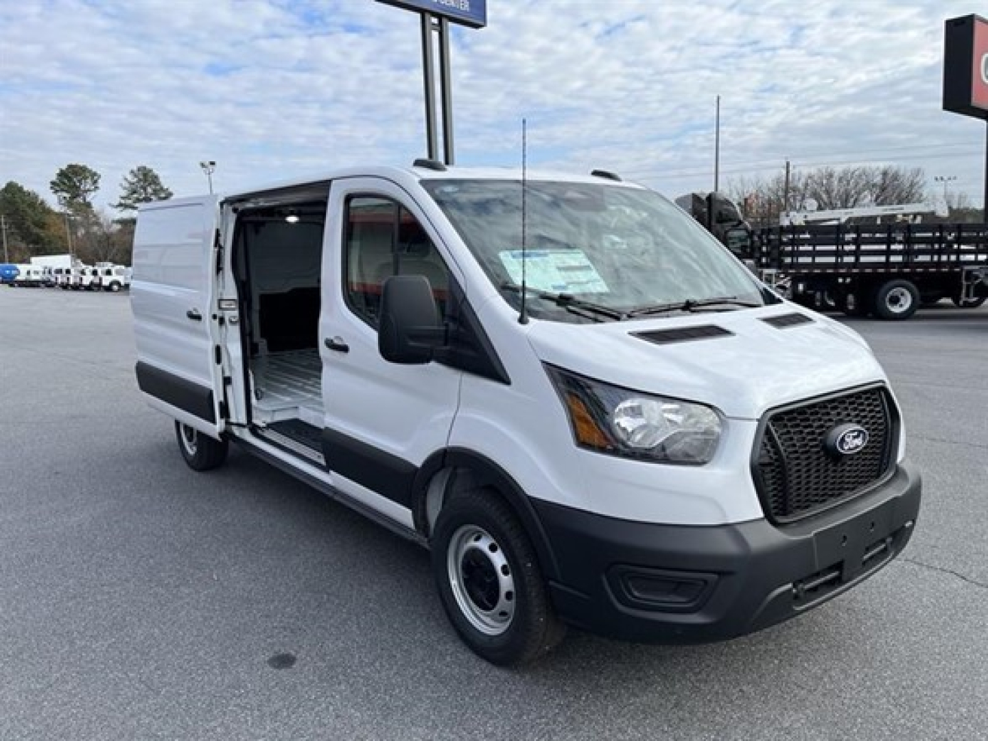 Ford Transit cargo van with side door open showing cargo area and front grille, white exterior, black bumper, in dealership lot.