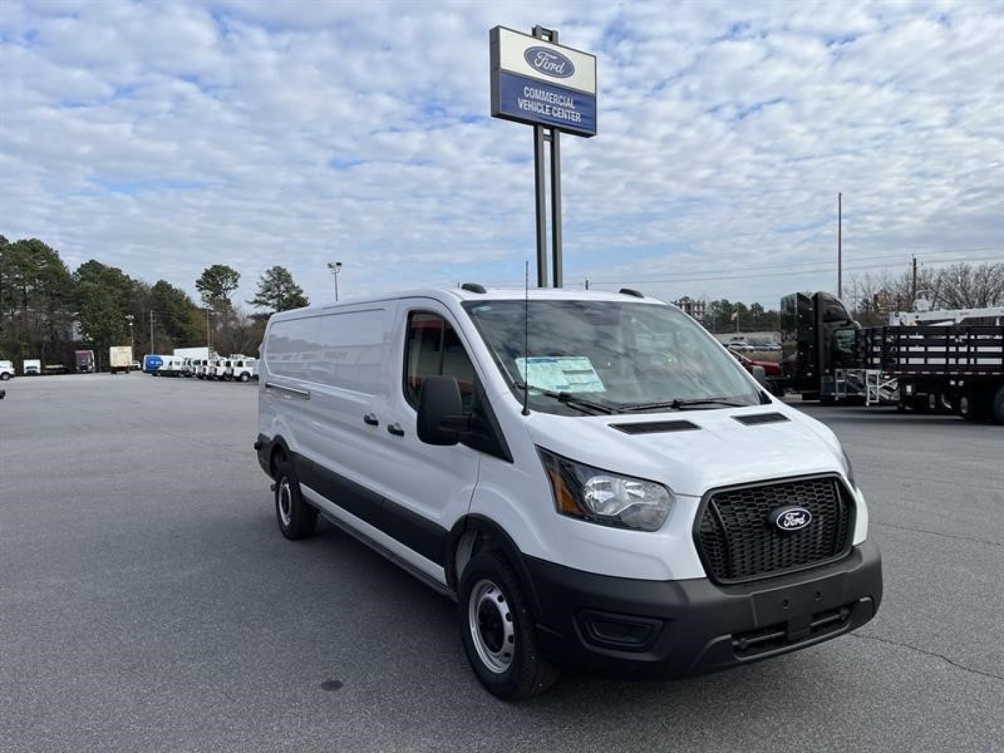 Ford Transit cargo van in a dealership lot showing front grille and side profile with stock sign in background.