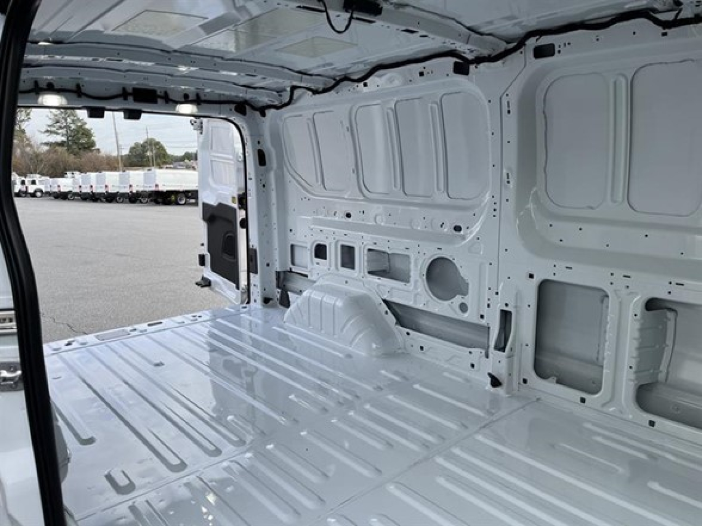 Ford Transit cargo van interior cargo area showing bare metal walls and floor with wheel well, rear doorway, and wiring channels.