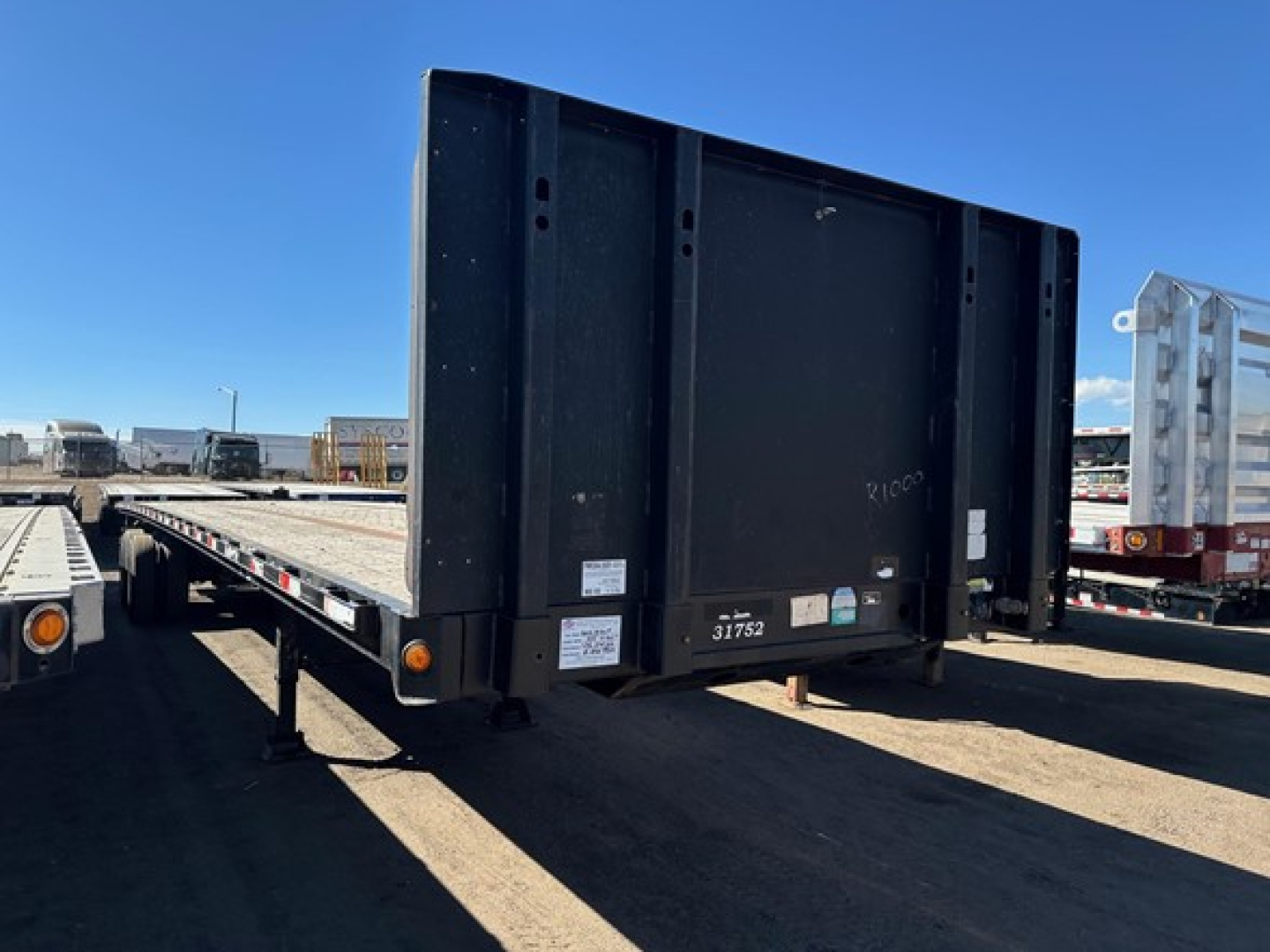Direct Steel flatbed trailer showing rear end with vertical risers and side rails, black steel exterior, tag stickers near hitch area, set on a dirt lot.