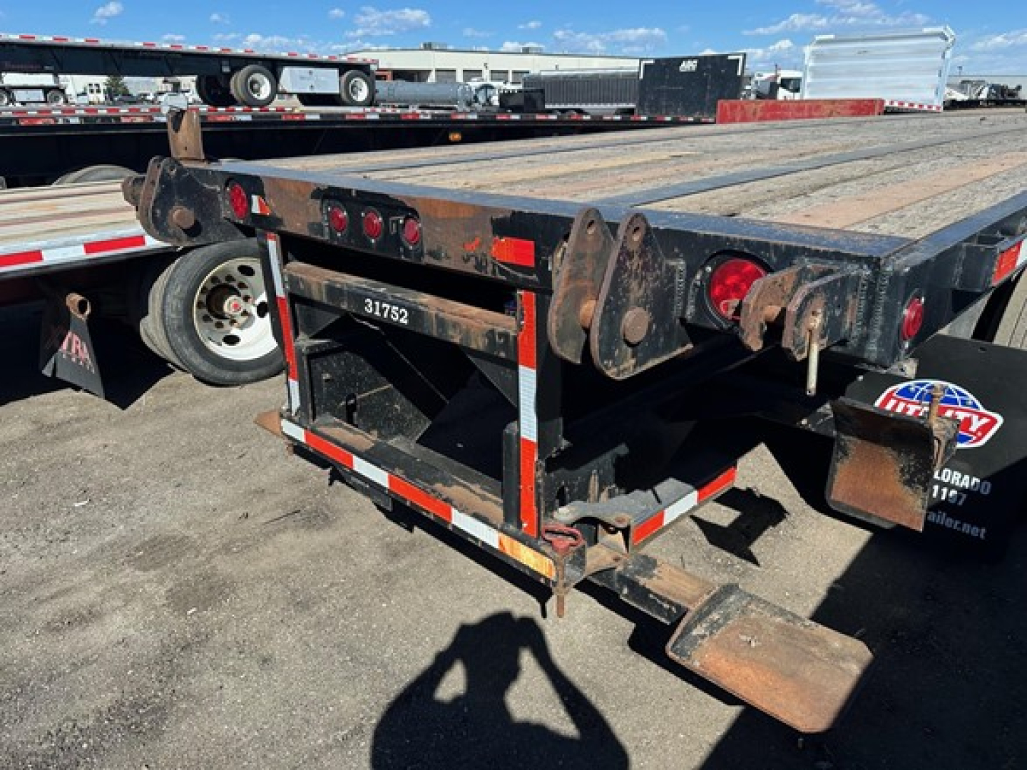Flatbed trailer rear view showing wooden deck, taillights, ramps and support structure; visible metal frame with rust accents and wheel with spare/dolly in background.