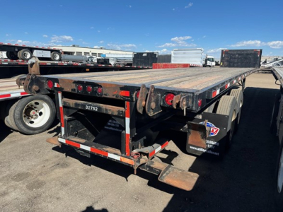 Flatbed trailer showing wooden deck with metal frame and dual rear axles; rear bumper and tail lights; some rust on hitch area; VIN/stock not visible in text on image.
