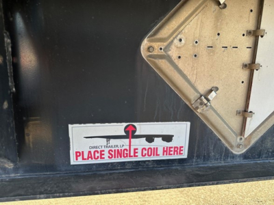 Sticker on trailer showing 'DIRECT TRAILER, LP PLACE SINGLE COIL HERE' with an arrow pointing to left and a hinge/metal plate nearby.