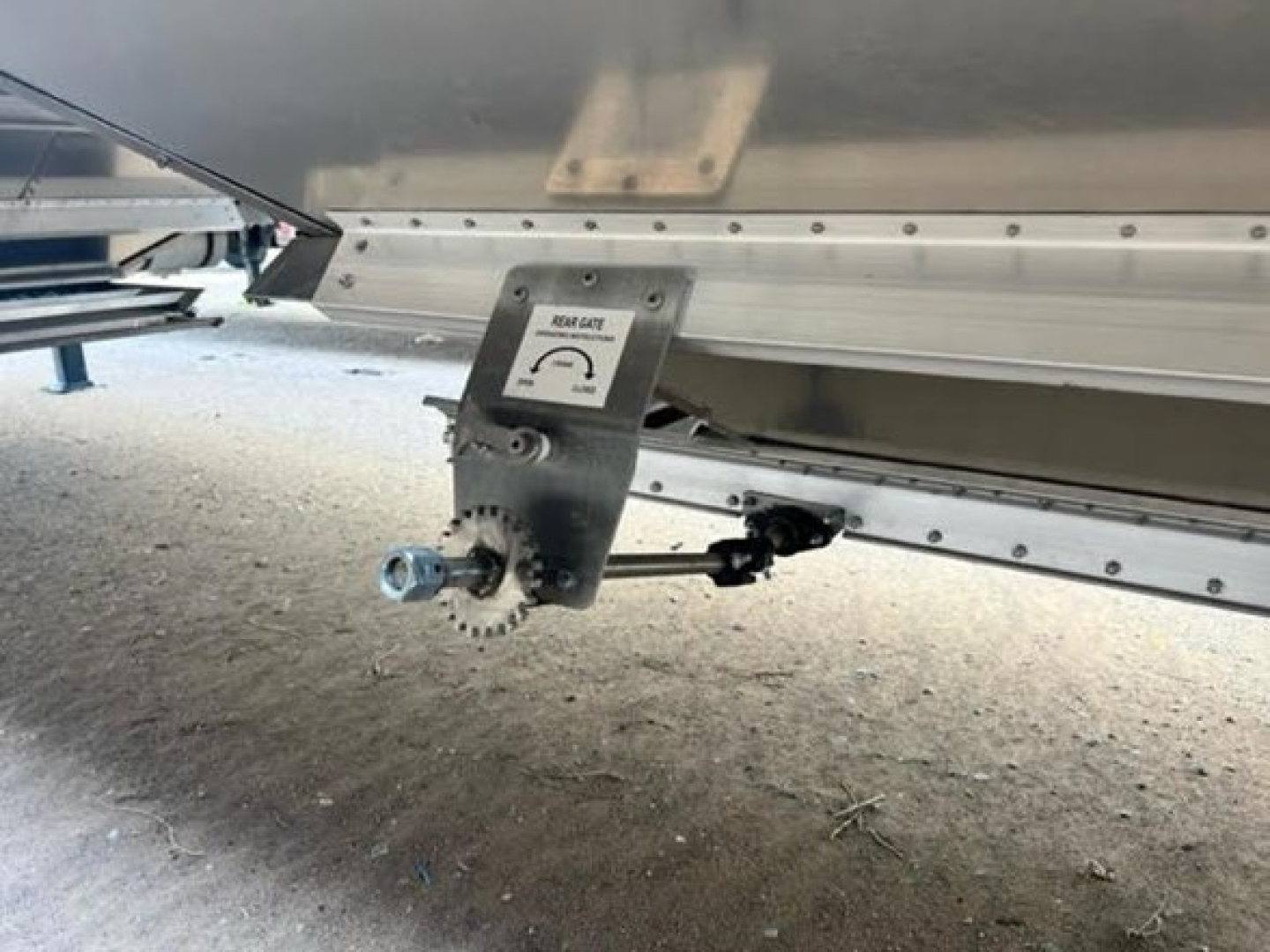 Trailer underside showing a metal hitch assembly with a gear-like sprocket and lockable pin, part of a Stoughton hopper trailer.