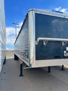 Trailer side view showing Stoughton Hopper trailer with blue front panel, riveted aluminum sides, front support legs, and a load chute. Visible text includes 'STOUGHTON' on the top front.
