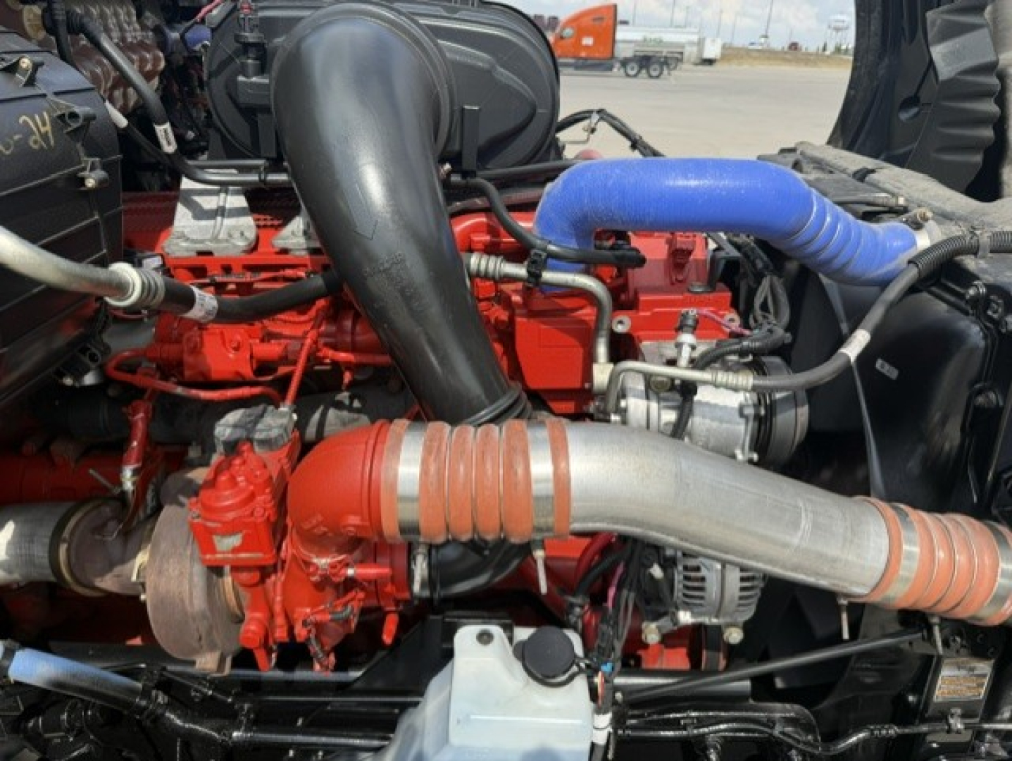 Kenworth T680 engine bay showing red engine components, large black intake, blue hose, and silver exhaust plumbing.