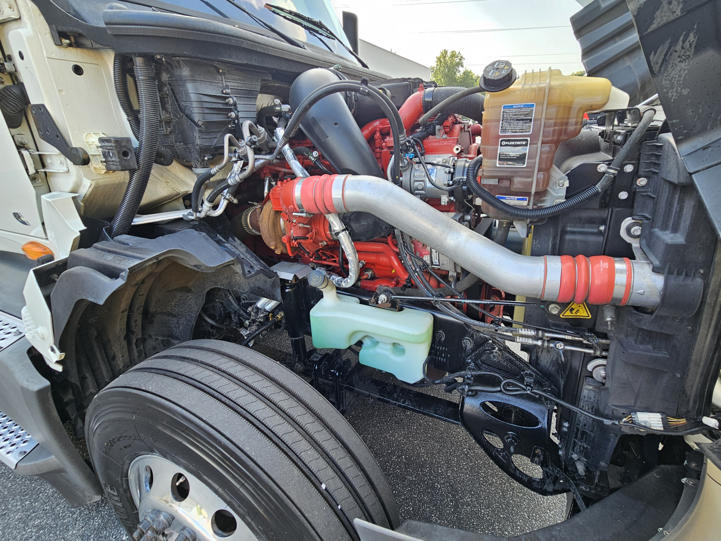 Truck engine bay of a 2021 International LT625 showing red engine, silver intake pipe, coolant reservoir, and various hoses. Visible components include coolant reservoir, front wheel, and engine block.