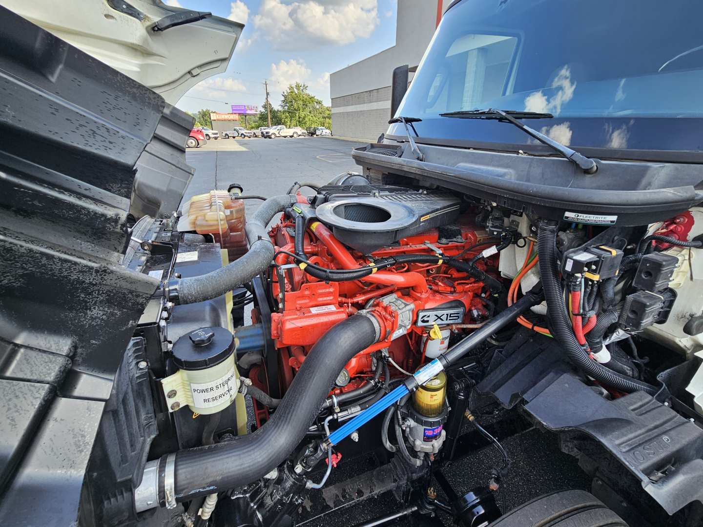 Truck engine bay showing Cummins/NT/Exis red engine with hoses, reservoirs, and air intake; front of cab visible with hood open