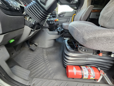 Truck cab interior showing driver's seat and dashboard with steering wheel, floor mat, and a red fire extinguisher on the floor near the seat; gear lever and control panels visible.