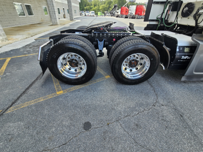 Dual rear axles of a semi-truck showing chrome wheels and mud flaps with part of sleeper cab; visible label on the tire reads 'Bridgestone' and accents of fuel lines and trailer hitch area.