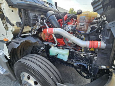 Truck engine bay of a 2021 International LT625 showing red engine, silver intake pipe, coolant reservoir, and various hoses. Visible components include coolant reservoir, front wheel, and engine block.