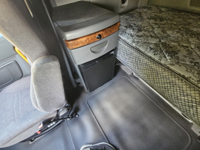 Interior cab view of a 2021 International LT625 truck showing a captain seat, storage drawer, wood trim, and a mattress in sleeper area.