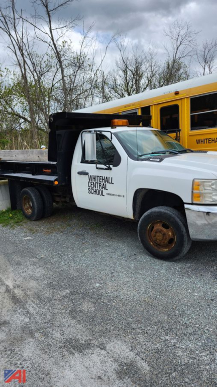 Truck front view showing white Chevrolet Silverado 3500HD dump bed with plow, school logo on door, and rusted wheel rims.