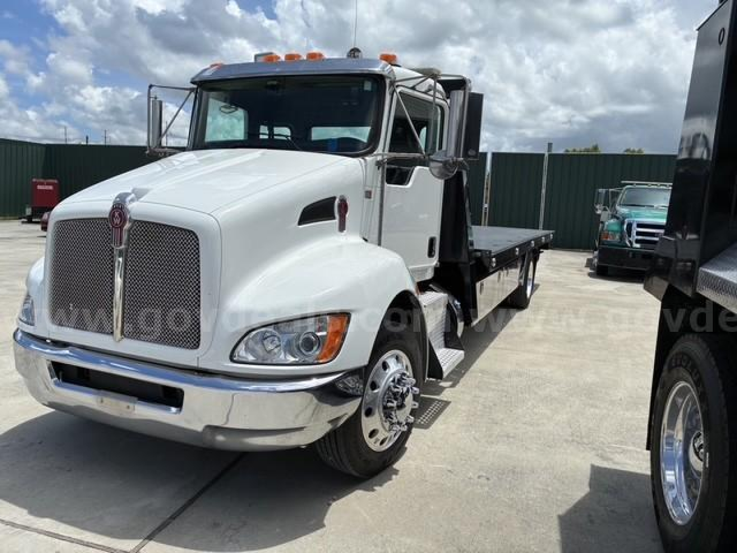 Kenworth T3 Series tow truck with flatbed showing front grille, headlights, and side mirror; white cab with chrome bumper and orange clearance lights.