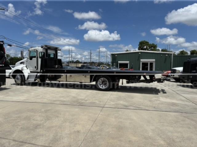 Kenworth T3 series tow truck with a long tandem flatbed, white cab, black flatbed, in a dealership lot.