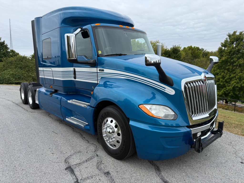 Blue 2024 International LT625 conventional sleeper truck with chrome grille and horizontal white stripes, front-right view showing fuel tanks and dual rear axles.