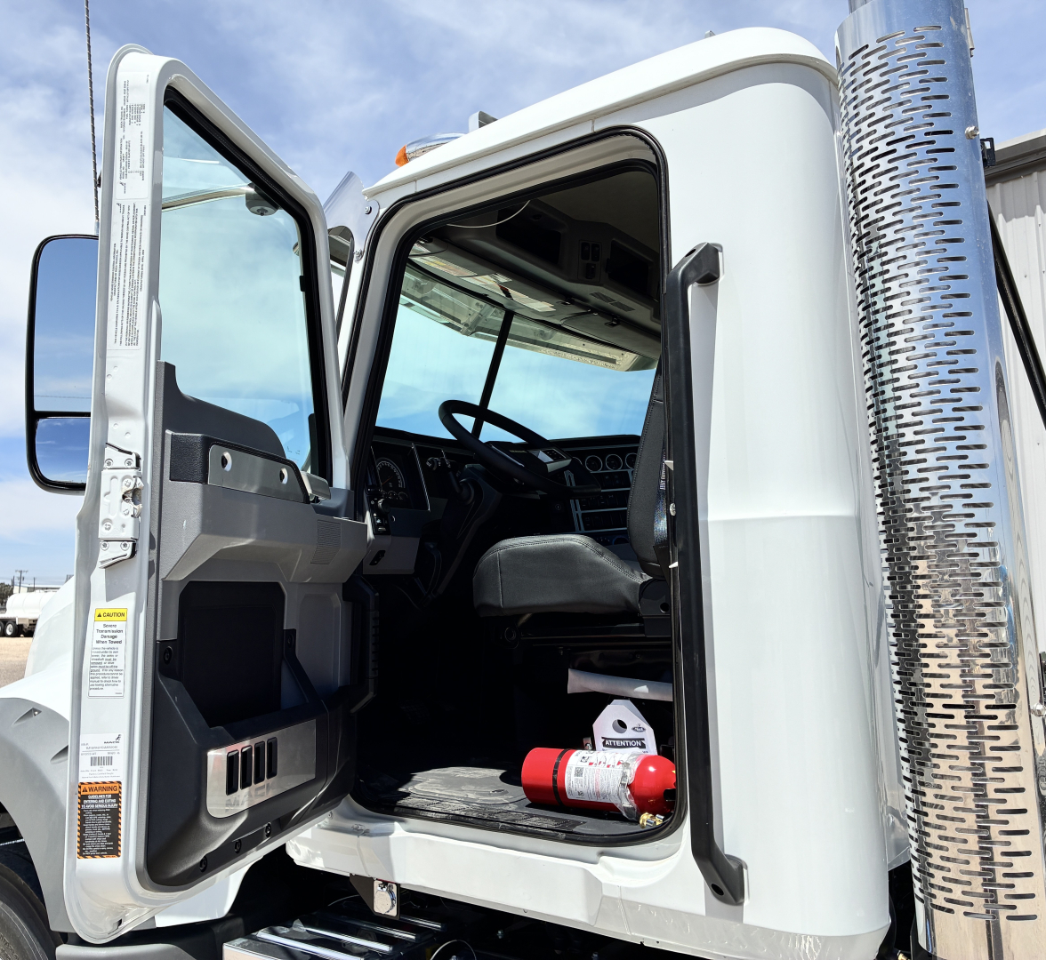 Truck cab interior and door area of a 2027 Mack GR64FT from the driver’s side with open door showing seat and dashboard; cylindrical chrome exhaust stack visible.