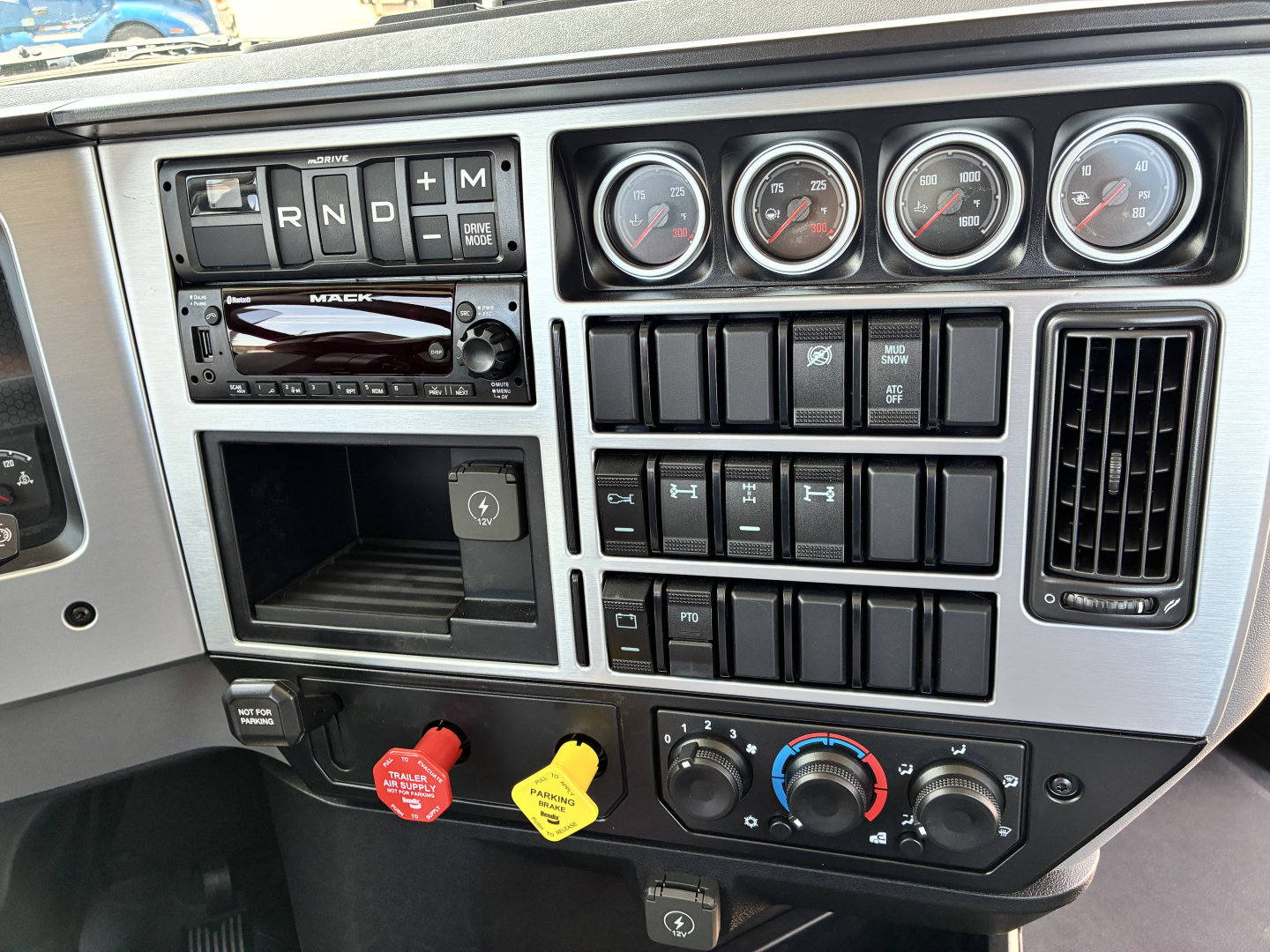 Dashboard controls and gauges of a 2027 Mack GR64FT truck showing transmission lever panel, multiple gauges, switches, and climate controls.
