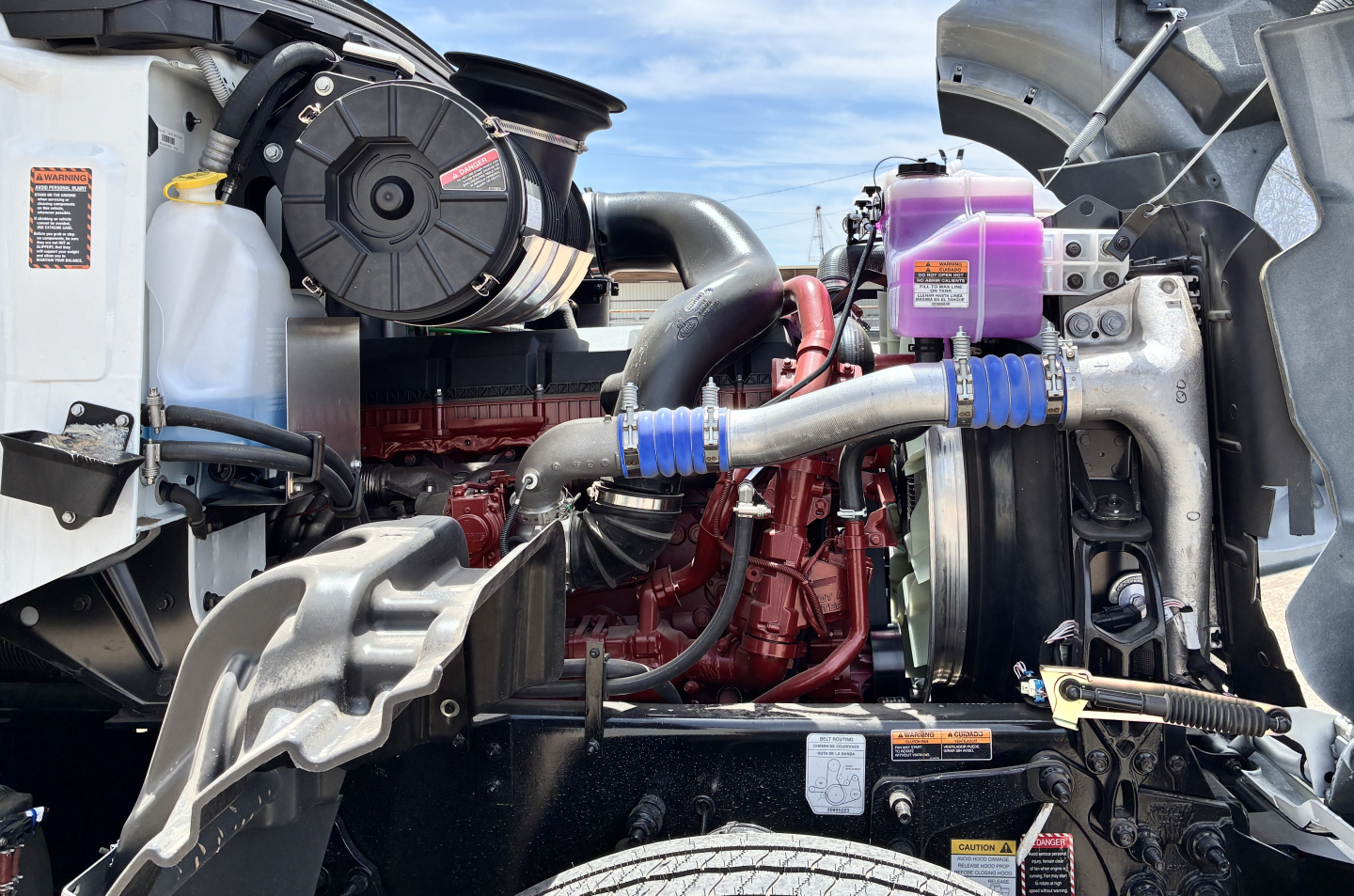 Mack GR64FT engine bay showing coolant reservoir, turbo intercooler piping, and red engine block with auxiliary components.
