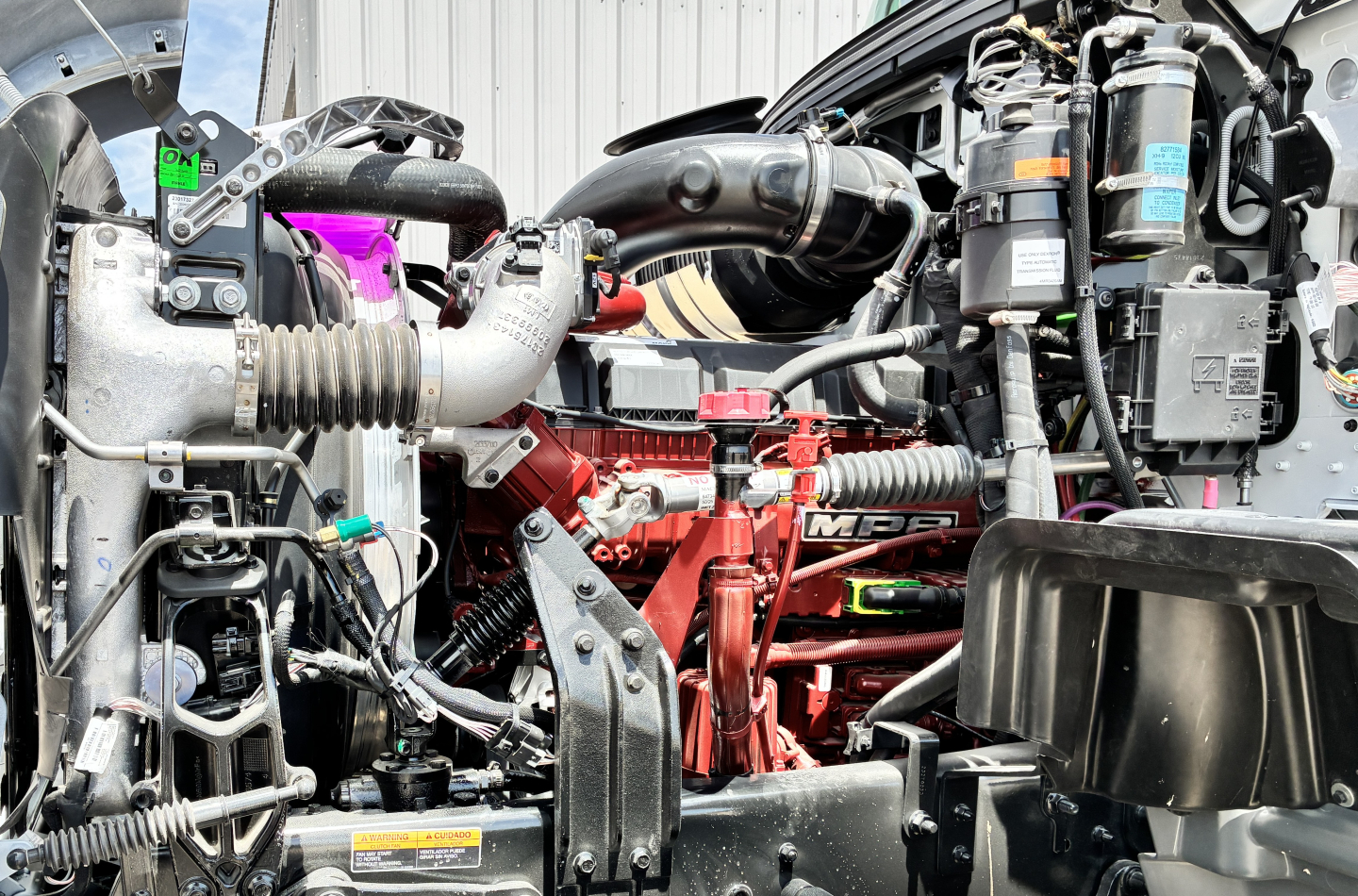 Engine bay of a 2027 Mack GR64FT showing red engine component, hoses, wiring, and various labels.