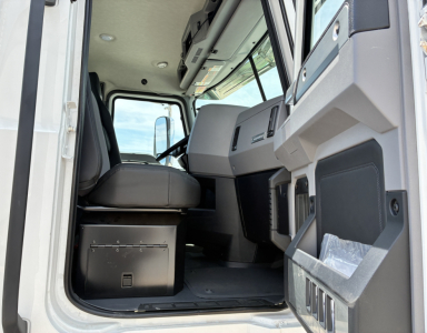 Truck cab interior in-view showing driver's seat, dashboard, and entry area of a new Mack GR64FT with light-gray and black trim.