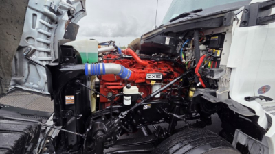 Kenworth T880 engine bay showing red Cummins X15 engine with blue rubber hose, white radiator area, and surrounding black chassis under-veed cab.
