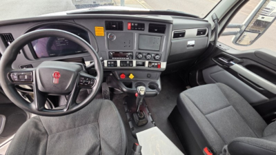 Truck cab interior showing steering wheel with Kenworth logo, dashboard with touchscreen display, numerous control buttons, manual gear shifter, and two front seats.