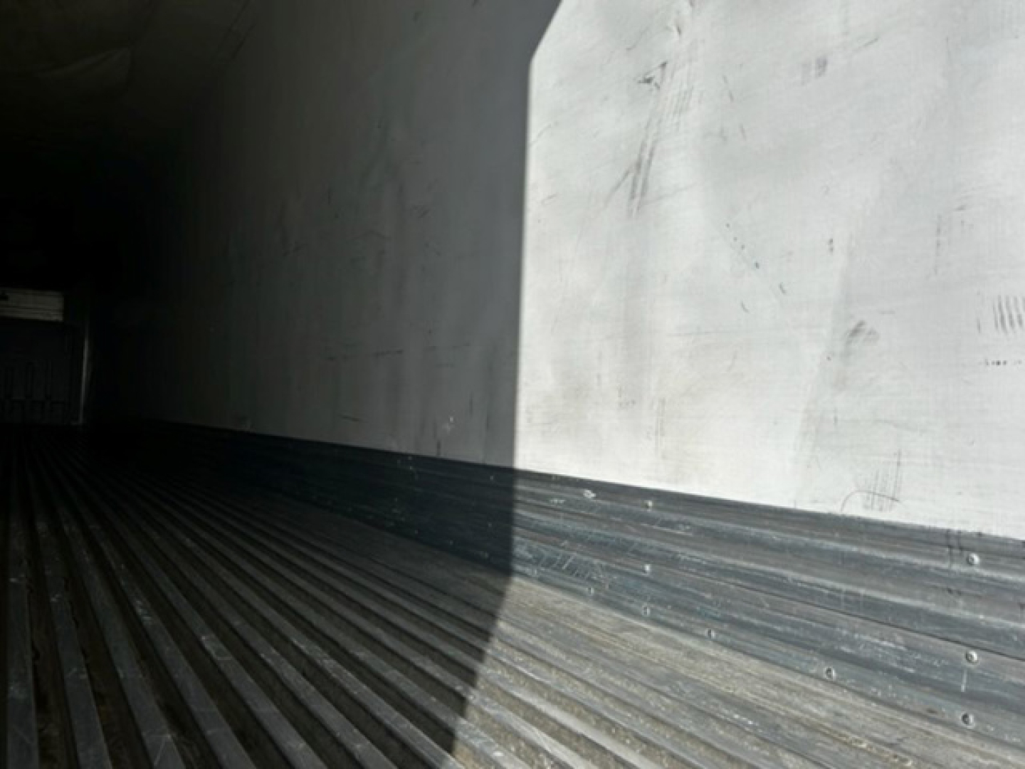 Trailer interior showing ribbed metal floor and a dirtied wall with light entering from the right, empty space inside a refrigerated trailer.