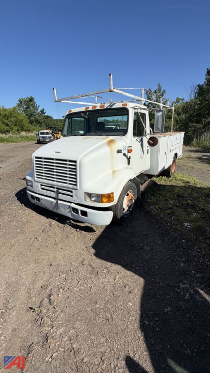 International 4700 LPX service truck with rack body, front view, white with rust stains and ladder rack on top.