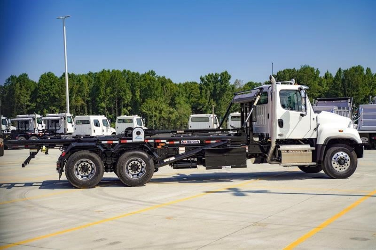 Freightliner 114SD roll-off truck, side view in a parking lot, long black flatbed with white cab and triple rear axles.