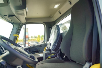Truck cab interior of a new Freightliner 114SD rolloff showing steering wheel, front seats, and plastic-covered passenger seat.