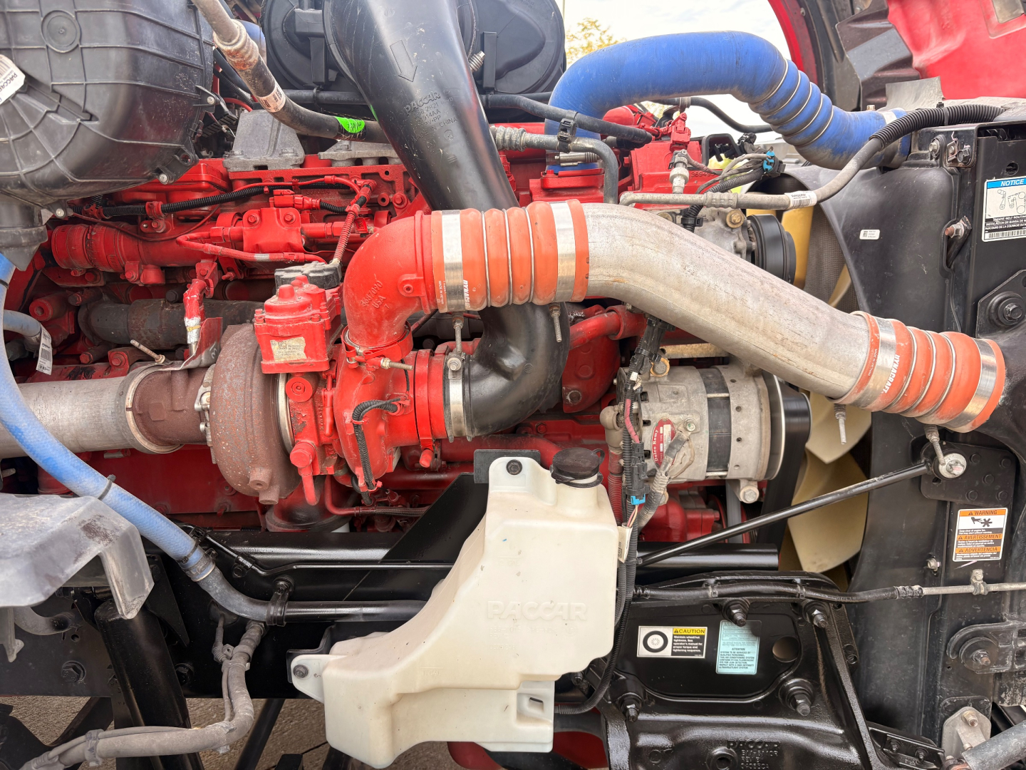 Kenworth T680 engine bay showing red turbocharger, large intake hose, coolant reservoir, and various blue hoses; visible engine block and surrounding components.