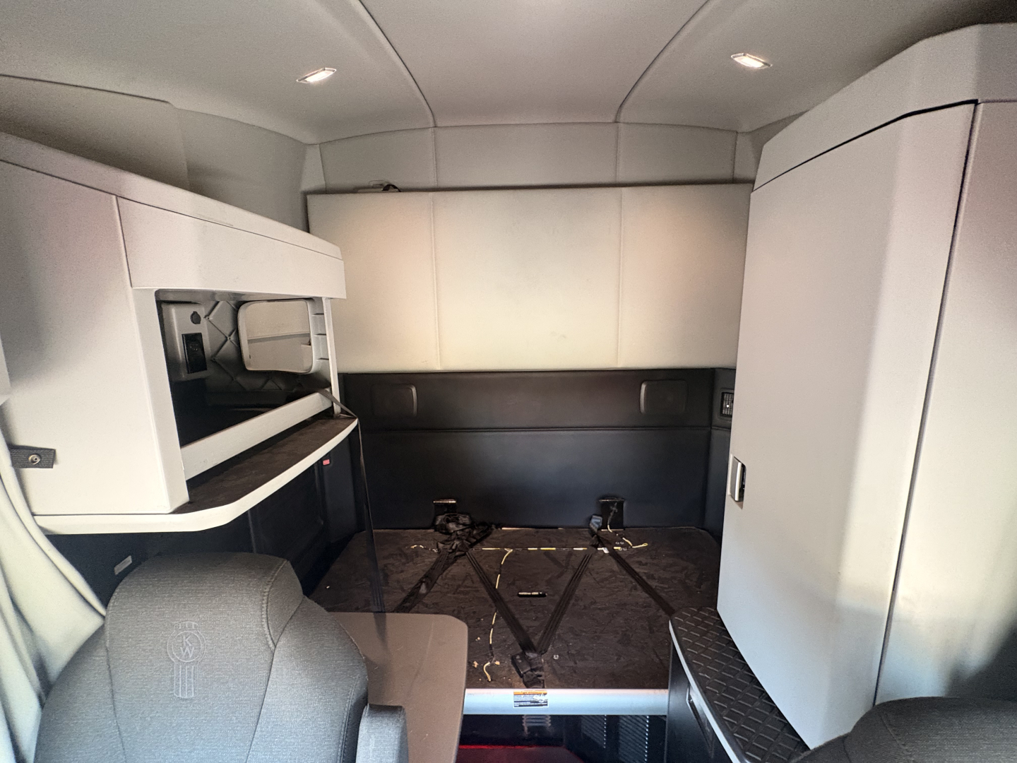 Interior cab view of a 2023 Kenworth T680 sleeper conventional truck showing workspace cabinet, storage lockers, and empty floor area.