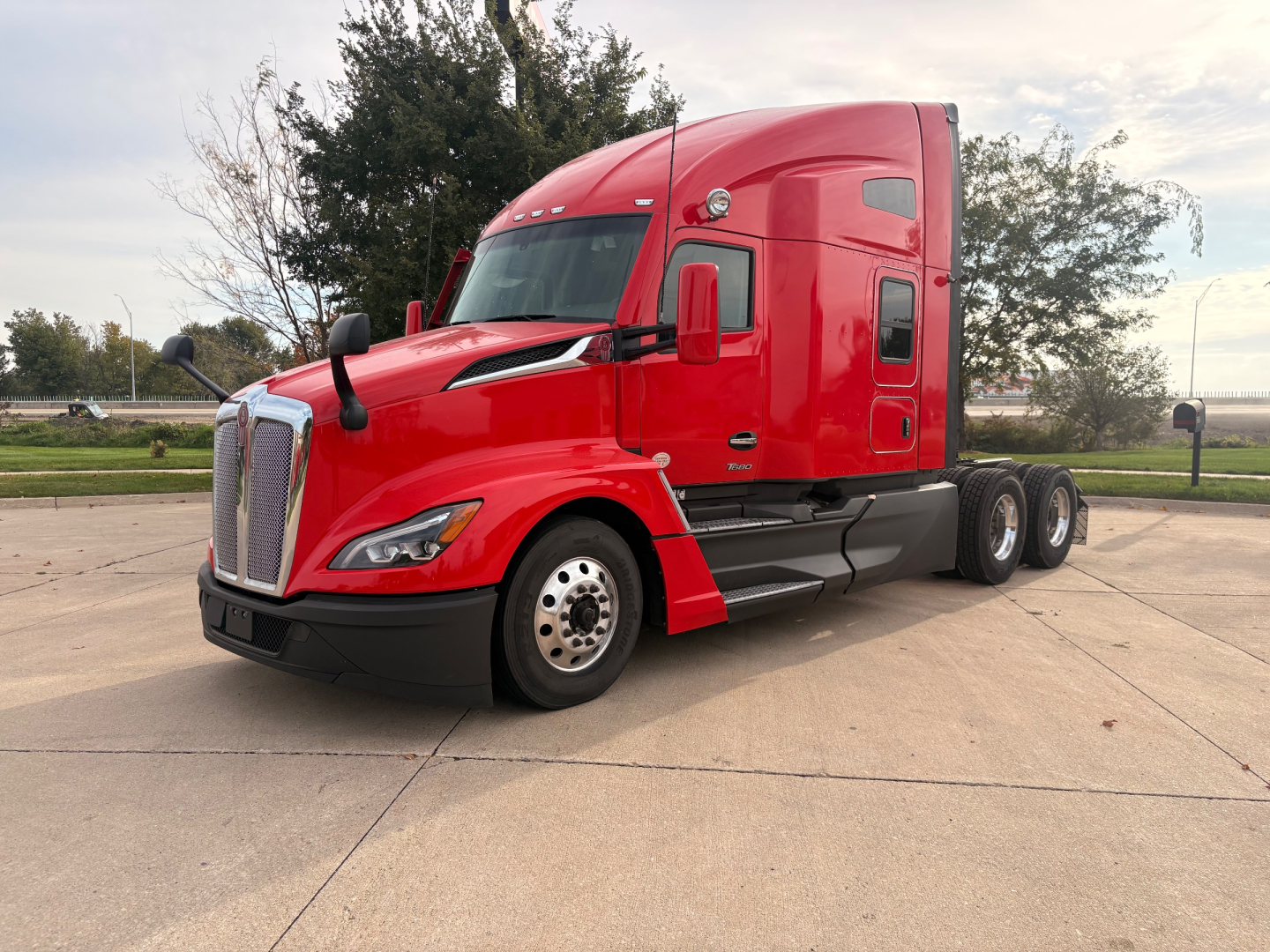 Kenworth T680 tractor trailer in red, front left view, with sleeper cab and three rear axles at the rear, no visible text on vehicle.