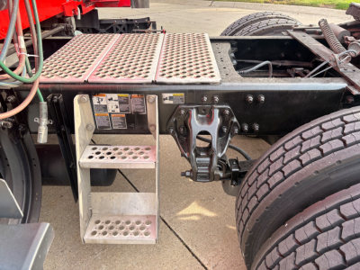 Truck chassis side rail with metal perforated platform steps and air/electrical lines near the cab mounting, visible wheels and frame of a Kenworth T680.