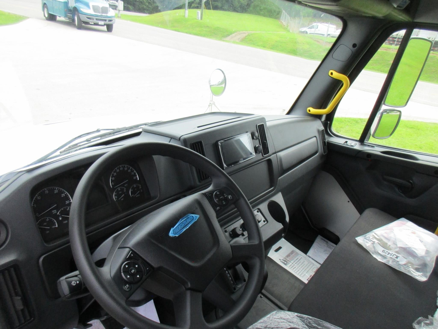 Dashboard and driver seat area of a Freightliner M2 106 cab interior showing steering wheel, instrument cluster, and center console in a new truck.