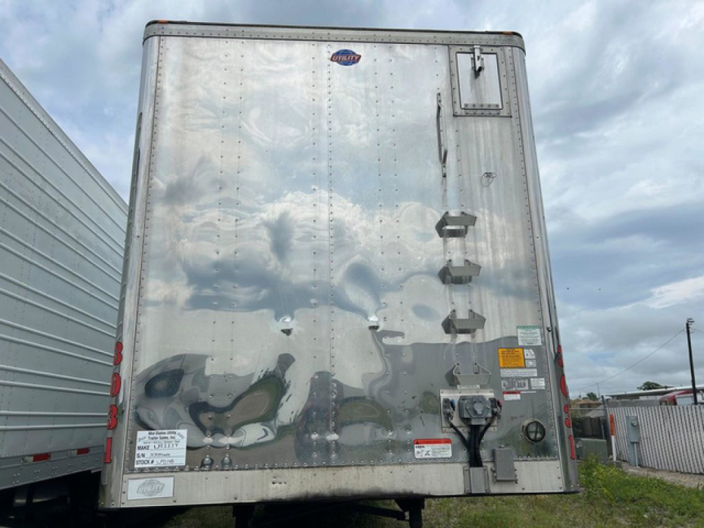 Back of a silver utility trailer showing rear doors with latches, hinges, reflectors, and bumper; stickers and a small lock mechanism near lower right.