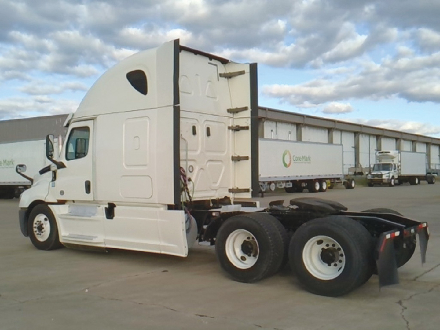 Freightliner Cascadia126 tractor-trailer in white, side view showing sleeper cab and rear wheels, with trailers in background.