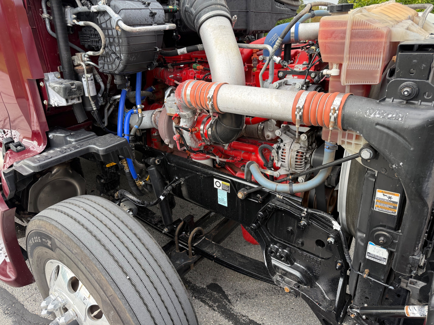Kenworth T680 engine bay view showing red engine, intercooler piping, hoses, and front suspension components.